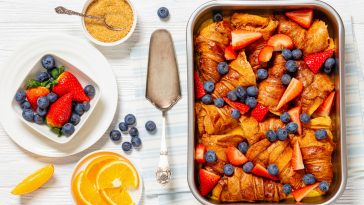 strawberry blueberry croissant casserole in baking dish on white wooden table with ingredients and cake shovel, horizontal view from above, flat lay