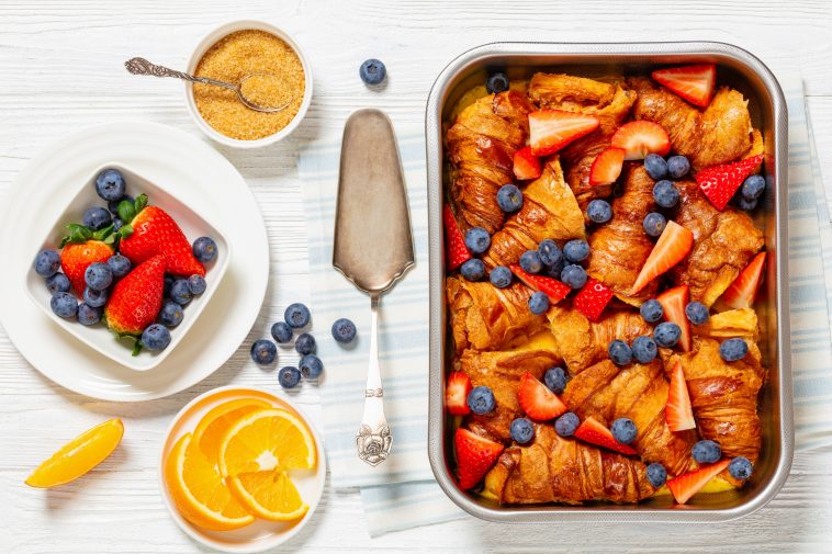 strawberry blueberry croissant casserole in baking dish on white wooden table with ingredients and cake shovel, horizontal view from above, flat lay