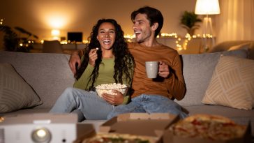 Cheerful Couple Using Home Cinema Projector Watching Comedy Movie Laughing, Eating Popcorn And Pizza Sitting On Sofa Indoors. Weekend Family Leisure, Entertainment And Fun