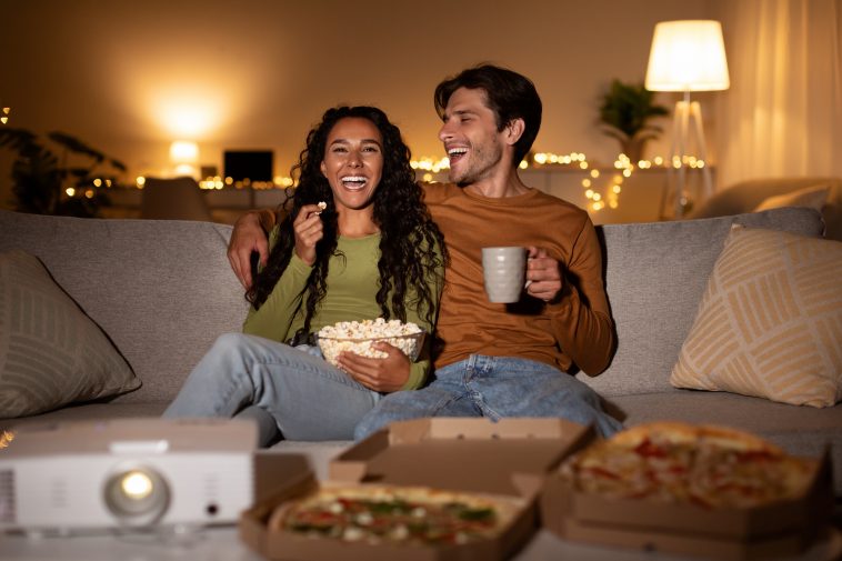 Cheerful Couple Using Home Cinema Projector Watching Comedy Movie Laughing, Eating Popcorn And Pizza Sitting On Sofa Indoors. Weekend Family Leisure, Entertainment And Fun