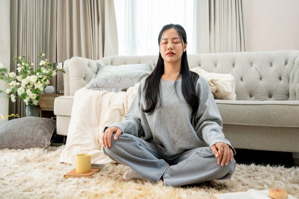 A relaxed, calm young Asian woman in comfy clothes is meditating in her living room in the morning. hobby, leisure, domestic life