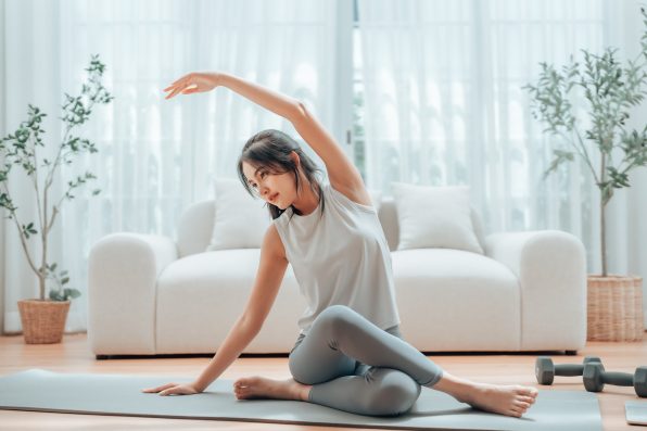 Happy young Asian woman practicing yoga and shoulder stretching at home sitting on floor in living room