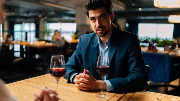 View from back of unrecognizable young woman to smiling elegant man in suit sitting at table with glasses of red wine having festive dinner at luxury restaurant. Happy loving couple enjoying talking.