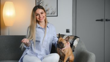 Young pretty woman in casual clothes hugging her dog sitting on the sofa in the living room of her cozy country house. Animal communication concept.