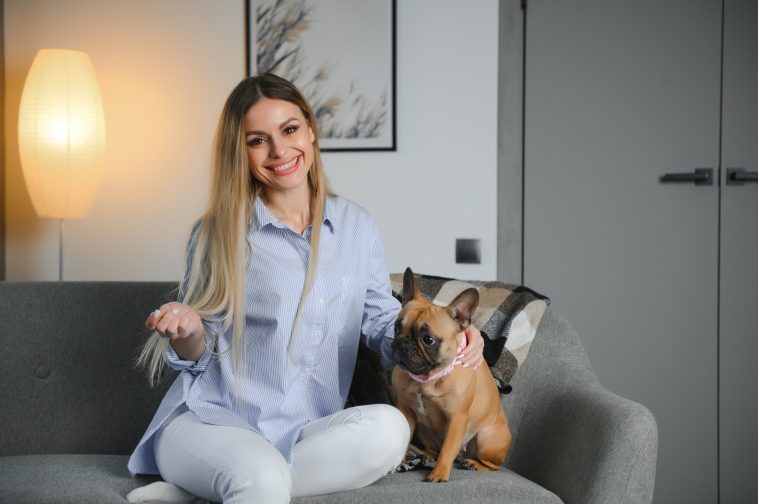 Young pretty woman in casual clothes hugging her dog sitting on the sofa in the living room of her cozy country house. Animal communication concept.