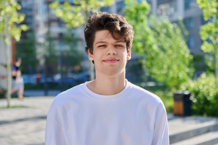 Headshot portrait of smiling, handsome student guy 19, 20 years old, looking at camera outdoors. Youth, urban style, young man, lifestyle concept