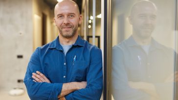 Waist up portrait of mature bald man smiling at camera while standing with arms crossed and posing confidently leaning against wall, copy space