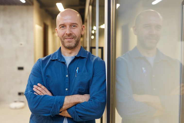 Waist up portrait of mature bald man smiling at camera while standing with arms crossed and posing confidently leaning against wall, copy space