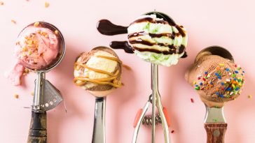 Variety of metal ice cream scoops with different ice cream and toppings on pink background.