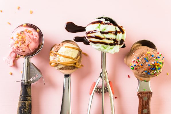 Variety of metal ice cream scoops with different ice cream and toppings on pink background.