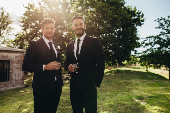 Portrait of handsome young groom standing with his friend having drinks at wedding party. Bridegroom and best men after wedding ceremony having drinks at park.