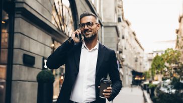 City walk. Technology. Coffee break. Business. Handsome man in suit is talking on the mobile phone, holding a cup of coffee and smiling while walking outdoors