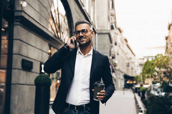 City walk. Technology. Coffee break. Business. Handsome man in suit is talking on the mobile phone, holding a cup of coffee and smiling while walking outdoors