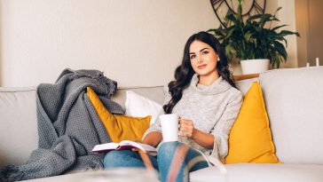 Young beautiful woman resting on a sofa, holding cup of tea or coffee, reading a book