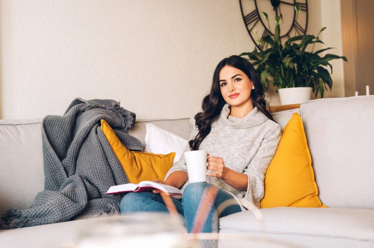 Young beautiful woman resting on a sofa, holding cup of tea or coffee, reading a book