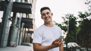 Happy hipster guy in casual t-shirt with copy space for brand label standing at urban setting and laughing during online communication via cellular gadget, positive cheerful man smiling outdoors