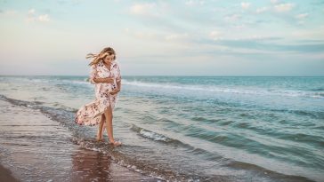 Beautiful happy young pregnant woman in a summer light dress hugging her belly and walks along the sea waves on a sunny summer evening during vacation. Natural Parenthood Concept. Copyspace