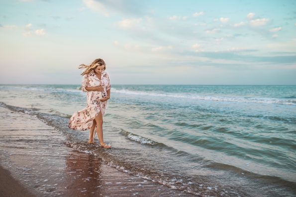 Beautiful happy young pregnant woman in a summer light dress hugging her belly and walks along the sea waves on a sunny summer evening during vacation. Natural Parenthood Concept. Copyspace