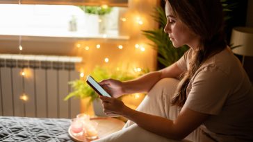 Pensive modern domestic woman in pajamas texting message smartphone sitting on bed with burning candles and illuminated garland. Relaxed female chatting mobile phone enjoy weekend leisure activity