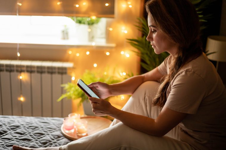 Pensive modern domestic woman in pajamas texting message smartphone sitting on bed with burning candles and illuminated garland. Relaxed female chatting mobile phone enjoy weekend leisure activity