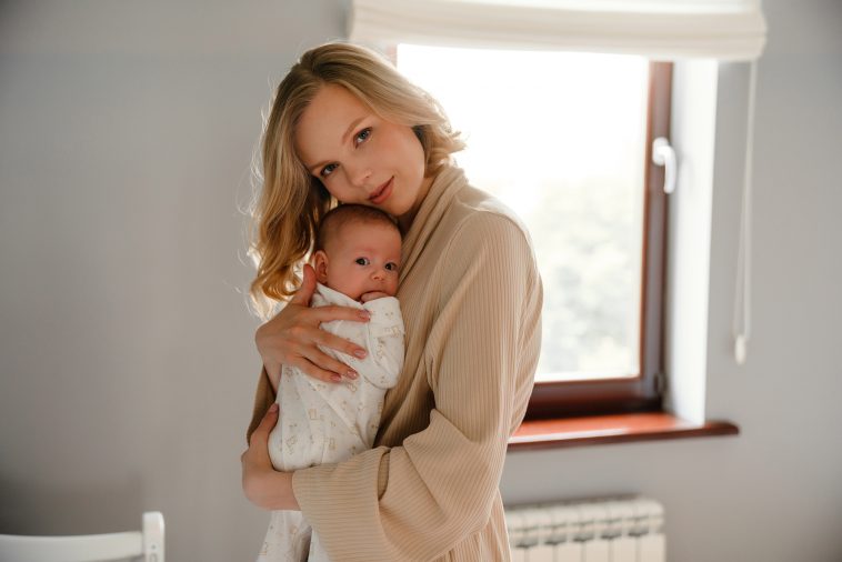 White young woman in housecoat holding her newborn baby in bedroom