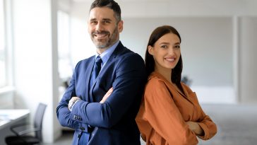 Two successful businesspeople standing back to back with arms crossed and smiling at camera, posing in modern office interior, copy space. Successful businessman and businesswoman