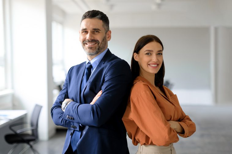 Two successful businesspeople standing back to back with arms crossed and smiling at camera, posing in modern office interior, copy space. Successful businessman and businesswoman