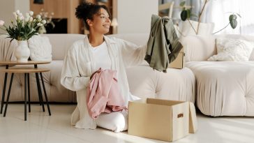 Young woman is smiling while unpacking clothes from a cardboard box in her living room