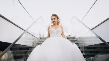 Beautiful young curly smiling blonde bride in white long dress walking down stairs in city outdoors. Wedding photography.