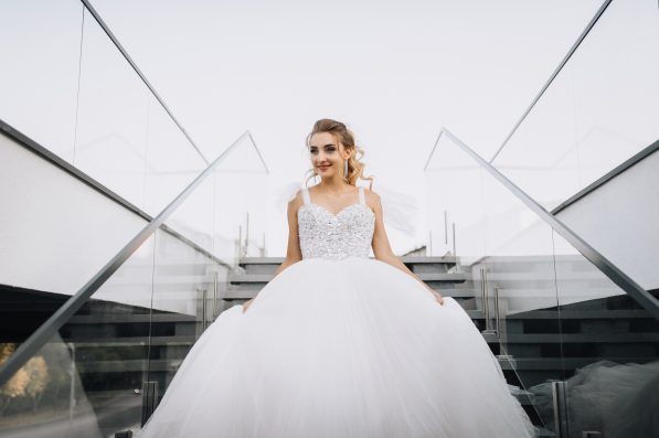 Beautiful young curly smiling blonde bride in white long dress walking down stairs in city outdoors. Wedding photography.