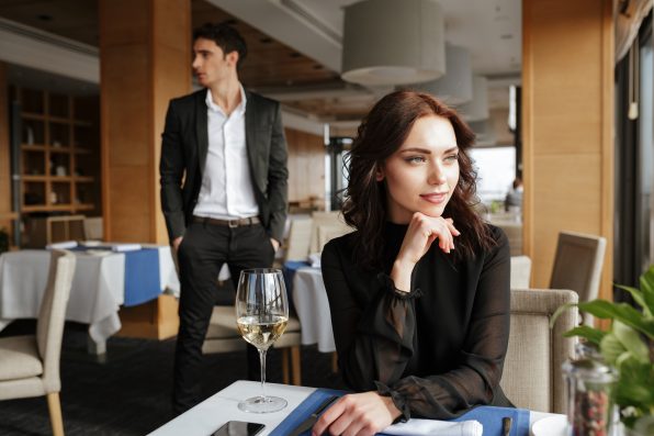 Woman in dress which sitting by the table in restaurant with man in suit on background