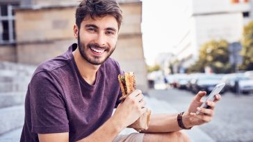 Young man eating lunch and using phone on stairs in the city