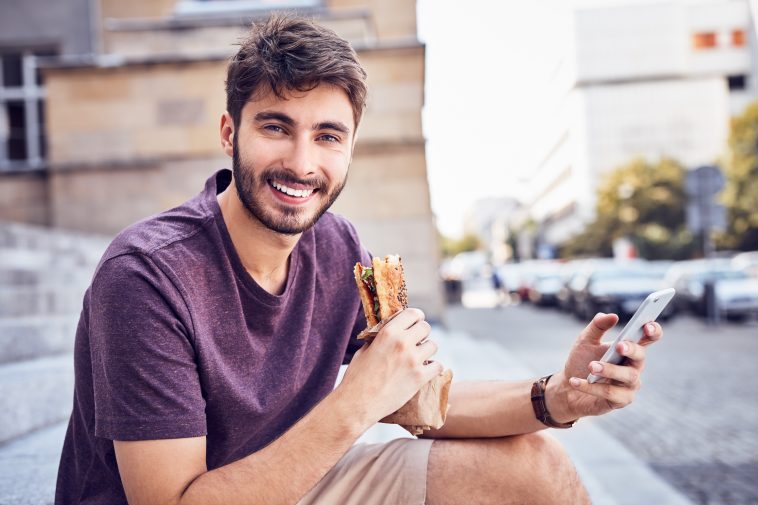 Young man eating lunch and using phone on stairs in the city