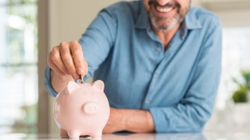 Middle age man save money on piggy bank with a happy face standing and smiling with a confident smile showing teeth