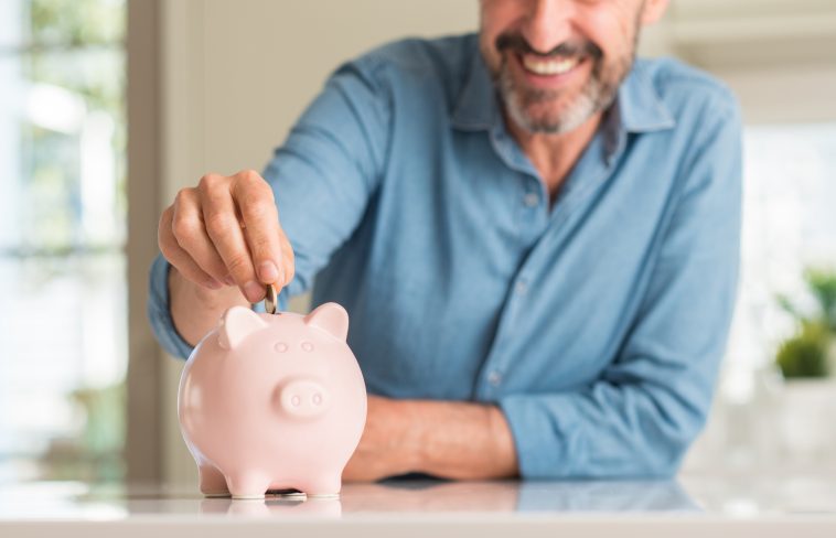 Middle age man save money on piggy bank with a happy face standing and smiling with a confident smile showing teeth