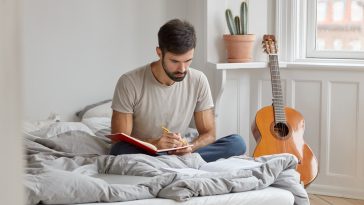 Photo of serious man with dark bristle, writes down story in diary about what happened during day, sits crossed legs at bed, wears casual clothing, has day off or weekend. Male records information