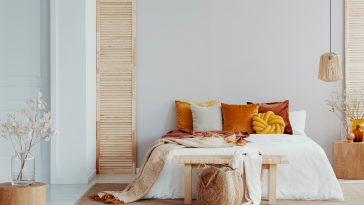 Brown and orange pillows on white bed in natural bedroom interior with wicker lamp and wooden bedside table with vase