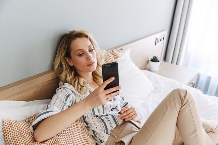 Beautiful young blonde woman relaxing in bed at home, using mobile phone