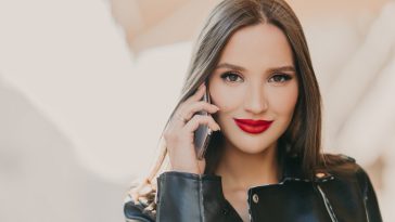 Close up shot of lovely woman talks with friend via mobile app, has makeup and red lips, dressed in leather jacket, has spare time outdoor. Online communication concept. Beauty and feminity.