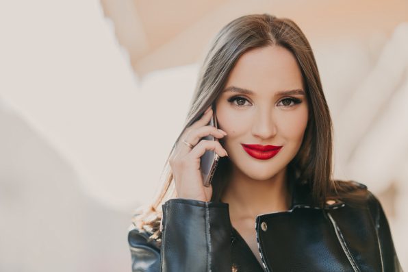 Close up shot of lovely woman talks with friend via mobile app, has makeup and red lips, dressed in leather jacket, has spare time outdoor. Online communication concept. Beauty and feminity.