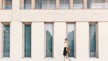 Business woman dressed in a leather pencil skirt, chiffon or silk blouse, wearing shoes with heels and sunglasses walking near a white building with tall windows.