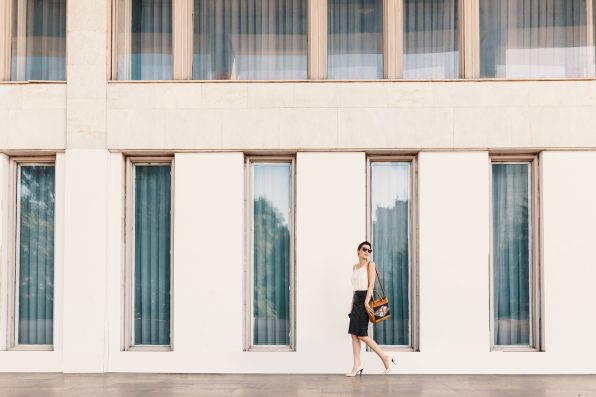 Business woman dressed in a leather pencil skirt, chiffon or silk blouse, wearing shoes with heels and sunglasses walking near a white building with tall windows.