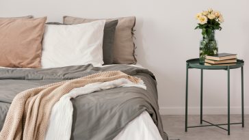 White roses in glass vase and books on stylish nightstand next to big bed with linen pillows and cozy blanket