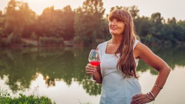 Mature woman enjoying glass of wine by river in autumn park. Senior woman admires landscape having drink