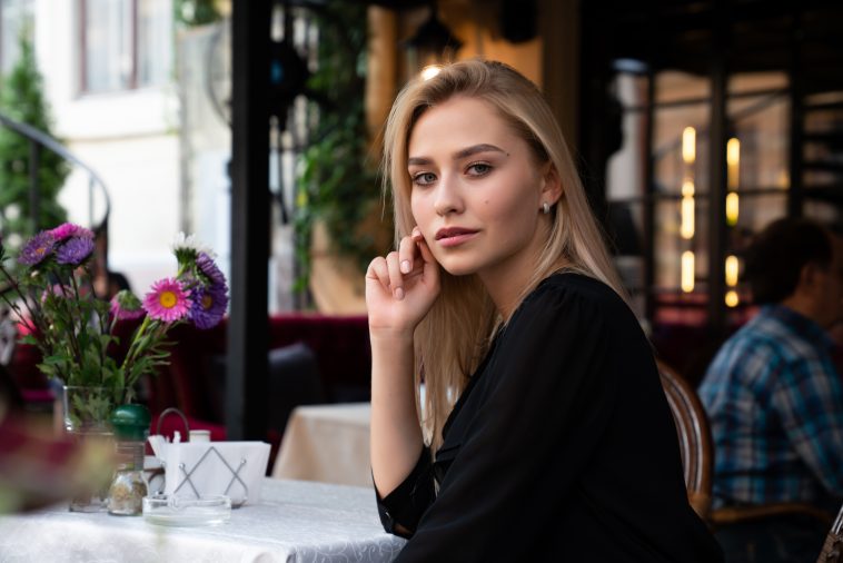 Mysterious blond girl in black dress sitting at a table in a cozy street cafe decorated with flowers and straightens hair with her hand, outdoor photo