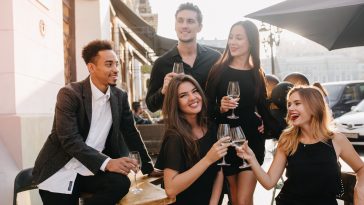 Joyful brunette man in black shirt looks away, holding glass of wine at party. Outdoor portrait of african guy in trendy jacket celebrating birthday with charming girls in cafe.