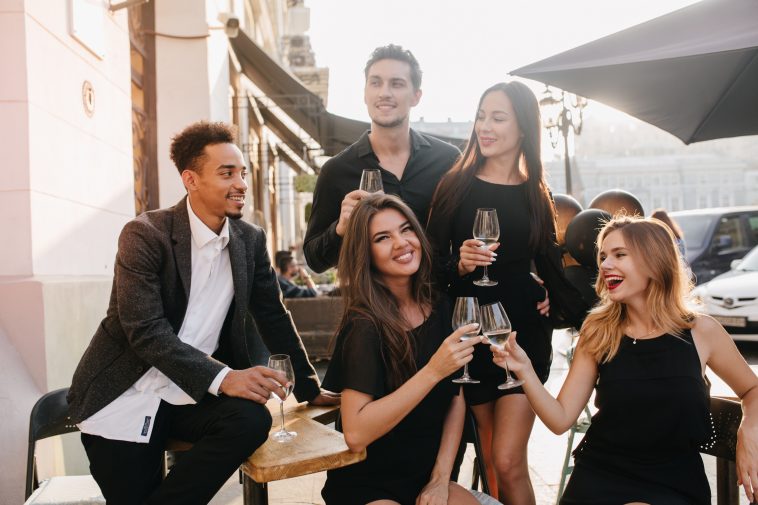 Joyful brunette man in black shirt looks away, holding glass of wine at party. Outdoor portrait of african guy in trendy jacket celebrating birthday with charming girls in cafe.