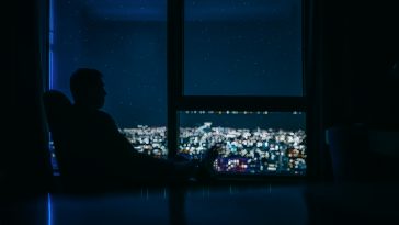 Silhouette of a young man on the panoramic window background with the glass of vine in her hand and is looking on the night city landscape with stars on the sky.