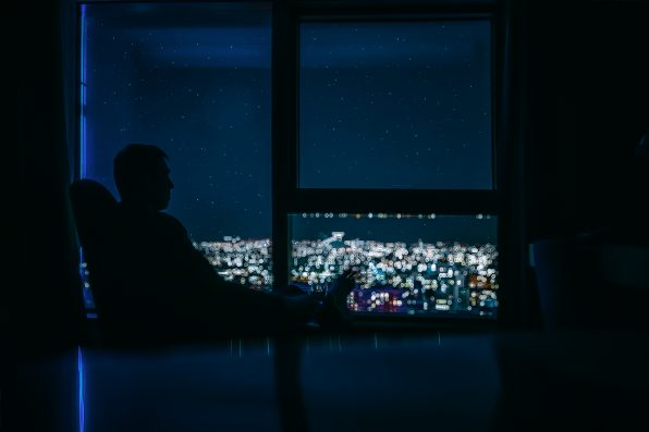 Silhouette of a young man on the panoramic window background with the glass of vine in her hand and is looking on the night city landscape with stars on the sky.