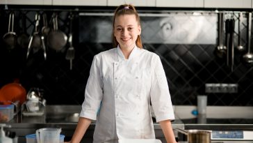 view on handsome young smiling woman chef wearing in white suit at kitchen. Blurred background